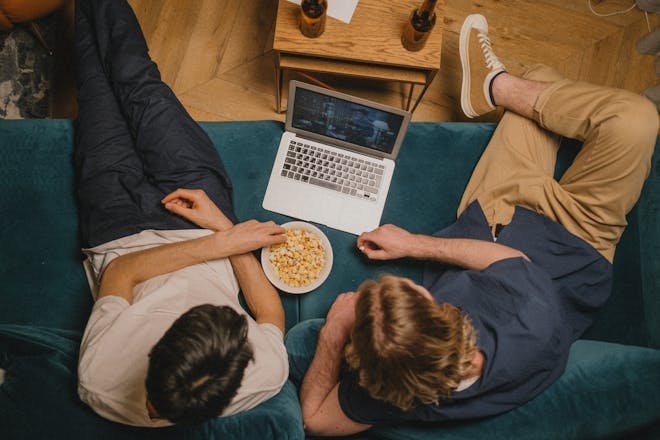 Two friends enjoying a ott movies on a laptop with popcorn from a cozy couch perspective.